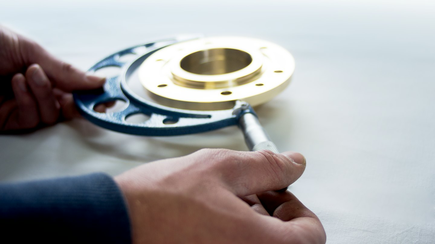 Hands use a blue micrometer to measure a golden metal part.